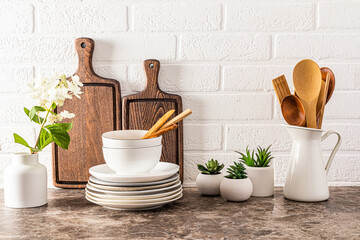 Fototapeta na wymiar A set of stylish kitchen utensils plates, bowls, cutting boards, spoons on a dark marble countertop opposite a white brick wall.