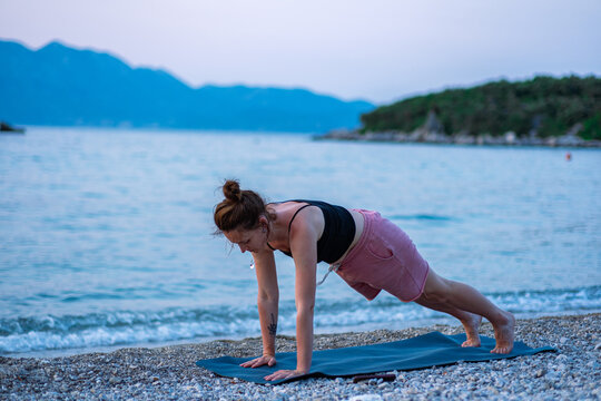 A young girl in a black tank top and pink shorts doing yoga on the sea beach. Yoga at sunset overlooking the beautiful Adriatic Sea and green shores. Photo promotes a healthy lifestyle, sports, yoga.
