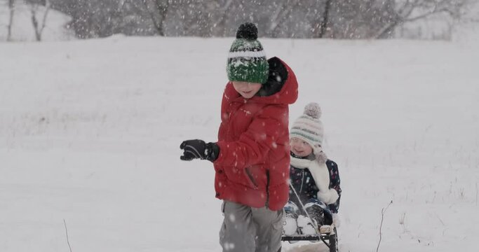 Boy pulls sleigh with small child across snow-covered field. Brother sleds younger sibling while it snows.