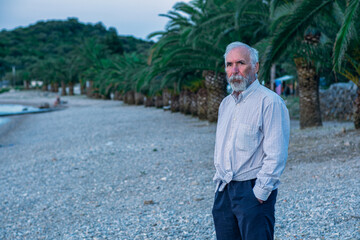 An old man on the beach in a white shirt and dark pants. An aged man with a beard walks alone on the beach at sunset. Portrait of a lonely gray-haired old man. Palm trees on the background.