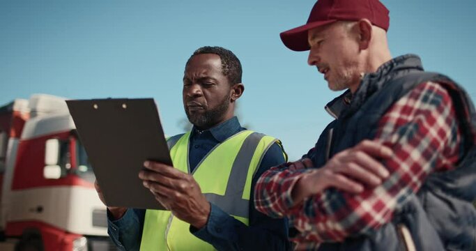 Hardworking mixed-race people discussing about upcoming import goods on their trucks. Busy manager with vest writing on clipboard paper some information about departure of trucks for delivery abroad.