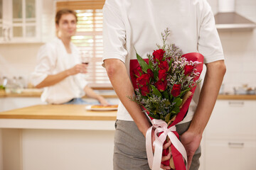 young gay couple looking at boyfriend and holding bouquet of roses behind his back for surprise in the kitchen