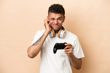 Fototapeta na wymiar Young Brazilian man playing with a video game controller isolated on beige background frustrated and covering ears