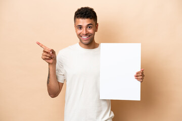 Young Brazilian man isolated on beige background holding an empty placard and pointing side