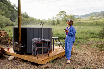 Naklejka premium Woman rests in a house in the mountains, heats up a hot tub for bathing and waiting with a smart phone. Concept of recreation on nature and hot bathing