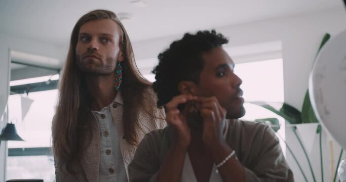 Young queer man staring at the mirror and arranging his earring with his partner next to him. 