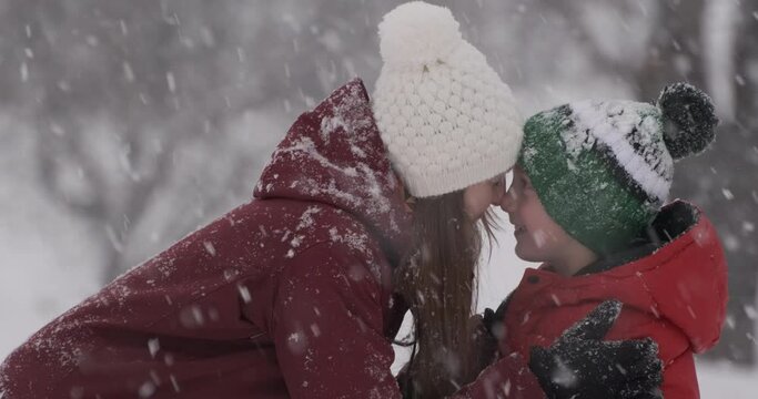 Mom and son rub their noses and laugh in snowy winter park. Happy young mother and child boy outdoors in winter day during snowfall.