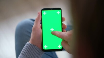 Young man sitting at home holding smartphone greenscreen mock-up in hand. Male person using chroma key mobile phone. Vertical mode. Touching, swiping display, tapping, surfing Internet social media