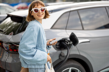Portrait of a young woman stands with a smart phone, waiting for her electric car to charge on a...