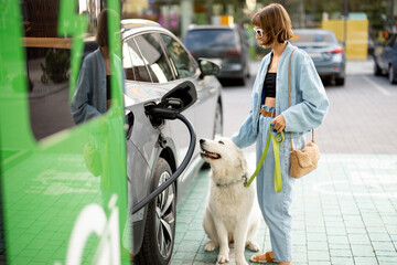 Happy woman with a huge white dog waiting for electric car to be charged on a public station...