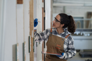 Portrait of happy smiling young female carpenter using digital tablet inspecting quality plank wooden at carpentry workshop. Happy smiling joiner female working in wood factory