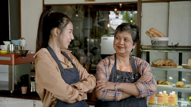 Senior mother with beautiful Asian daughter, standing in front checkout counter taking orders inside cafe, which is its own bakery, stand crossed arms look at camera take pictures promote shop.
