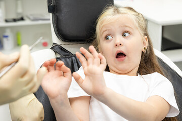 Childish fear. Cute little girl sitting in the dentist chair and covering her mouth in fear, being afraid of a dental drill in the hands of her doctor