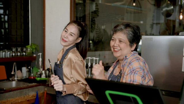 Elderly single mother with beautiful Asian daughter happily dances behind checkout counter taking orders in own small business cafe. after closing sales from many applications which is its own bakery