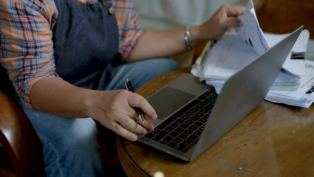 Close-up hands elderly woman is mother, holding pen, opened stack documents lying on table, finished opening document, woman turned around typed account information into laptop computer.