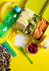 School lunch box with sandwiches, carrot sticks, apple, banana, lettuce, hummus and raspberries. Healthy school lunch concept. Yellow background and note with text - have a nice day mom. Top view 