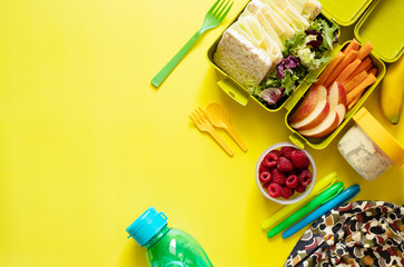 School lunch box with sandwiches, carrot sticks, apple, banana, lettuce, hummus and raspberries. Healthy school lunch concept. Yellow background. Top view. Place for text.