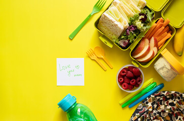 School lunch box with sandwiches, carrot sticks, apple, banana, lettuce, hummus and raspberries. Healthy school lunch concept. Yellow background and note with text - love you mom. Top view 