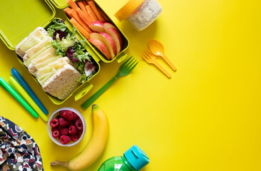 Obraz na płótnie Canvas School lunch box with sandwiches, carrot sticks, apple, banana, lettuce, hummus and raspberries. Healthy school lunch concept. Yellow background. Top view. Place for text.