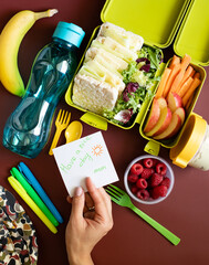 School lunch box with sandwiches, carrot sticks, apple, banana, lettuce, hummus and raspberries. Healthy school lunch concept. Female hand holding a note with text - have a nice day mom. Top view