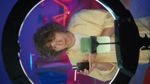 Young man vlogger in casual clothing advertises a perfume in front of his smartphone at home studio. Vertical video