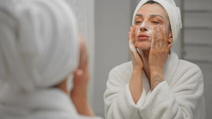 Female close up beauty care portrait. Adult woman wearing towel on head using moisturizing foam for face washing, morning skincare routine