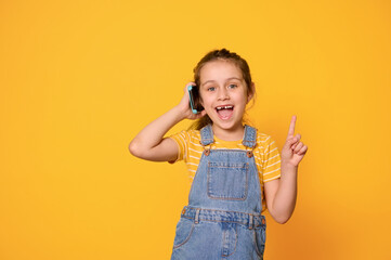 Beautiful amazed little kid girl talking on smart mobile phone, pointing finger aside a copy ad space, yellow background