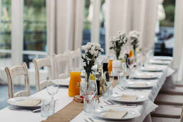 Reserved table for a wedding. Table setting in white moans. Transparent vases with wild flowers on the table. White tablecloth and cutlery. Orange juice on the table.