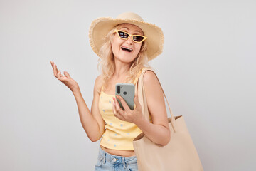 Portrait positive mature blond woman in straw hat, yellow top and sunglasses holding smartphone and shopping bag isolated on white studio background