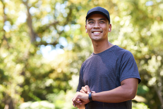 Attractive Asian male syncs fitness device with earphones, running in leafy park