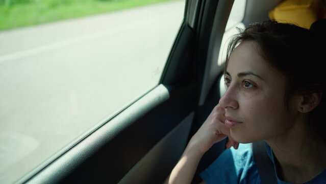 young woman sitting close to window in moving car and looking outside. girl is bored during long road trip in summer in car. summertime vacation car traveling