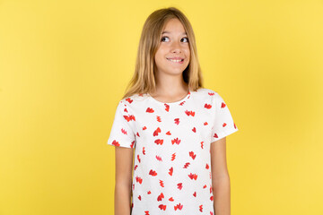 Obraz premium Amazed blonde kid girl wearing polka dot shirt over yellow studio background bitting lip and looking tricky to empty space.