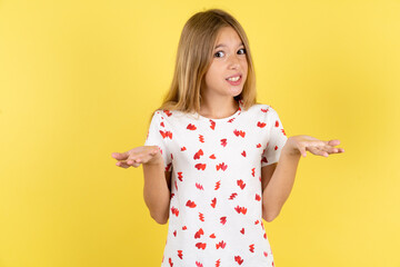 Clueless blonde kid girl wearing polka dot shirt over yellow studio background shrugs shoulders...