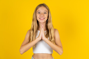 blonde kid girl wearing white T-shirt over yellow studio background praying with hands together asking for forgiveness smiling confident.
