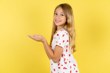 blonde kid girl wearing polka dot shirt over yellow studio background pointing aside with hands...
