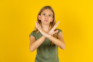 blonde kid girl wearing green T-shirt over yellow studio background Rejection expression crossing arms doing negative sign, angry face