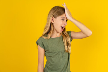 blonde kid girl wearing green T-shirt over yellow studio background surprised with hand on head for mistake, remember error. Forgot, bad memory concept.
