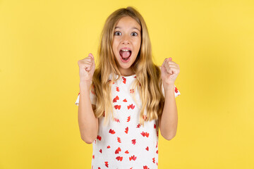 blonde kid girl wearing polka dot shirt over yellow studio background celebrating surprised and...