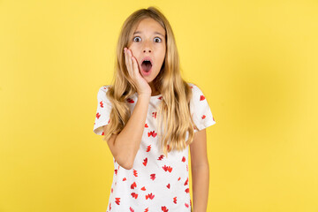 Shocked blonde kid girl wearing polka dot shirt over yellow studio background looks with great...