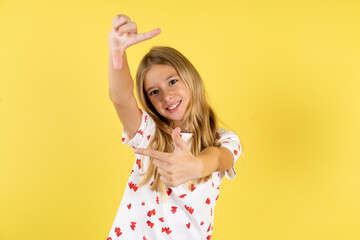 blonde kid girl wearing polka dot shirt over yellow studio background  making finger frame with...