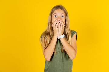 Vivacious blonde kid girl wearing green T-shirt over yellow studio background , giggles joyfully, covers mouth, has natural laughter, hears positive story or funny anecdote - obrazy, fototapety, plakaty