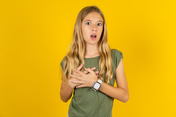 Scared blonde kid girl wearing green T-shirt over yellow studio looks with frightened expression, keeps hands on chest, being puzzled to notice something strange, People, hush reaction and emotions.