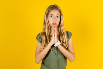 Positive blonde kid girl wearing green T-shirt over yellow studio background smiles happily, glad...