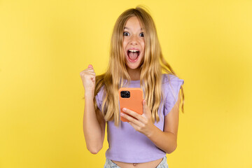Portrait of blonde kid girl wearing violet T-shirt over yellow studio background holding in hands...