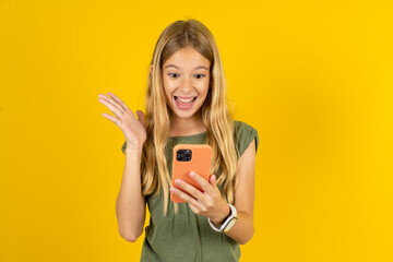 blonde kid girl wearing green T-shirt over yellow studio background  holding in hands cell reading...