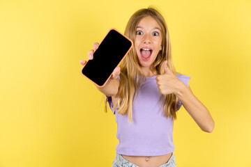 Portrait blonde kid girl wearing violet T-shirt over yellow studio background holding in hands cell...