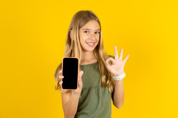 blonde kid girl wearing green T-shirt over yellow studio background holding in hands cell showing...