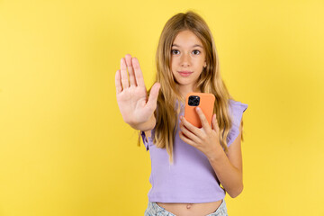 blonde kid girl wearing violet T-shirt over yellow studio background using and texting with smartphone with open hand doing stop sign with serious and confident expression, defense gesture