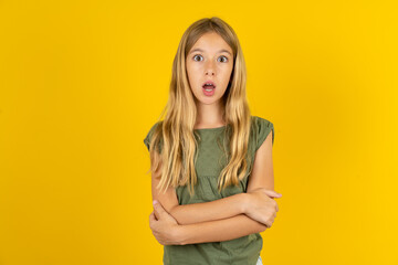 Shocked embarrassed blonde kid girl wearing green T-shirt over yellow studio background keeps mouth...