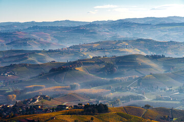 Wide panorama of a valley in Piedmont, Italy, with sunlit vineyards in the foreground, and most of the image space occupied by successive mountain chains emerging from the early morning mist